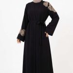 Mannequin portant une abaya noire élégante avec des broderies florales dorées sur l'épaule et les manches, collection Orkidia Fashion.