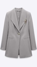 Blazer gris cintré élégant avec broche chaîne dorée, parfait pour une tenue professionnelle femme.